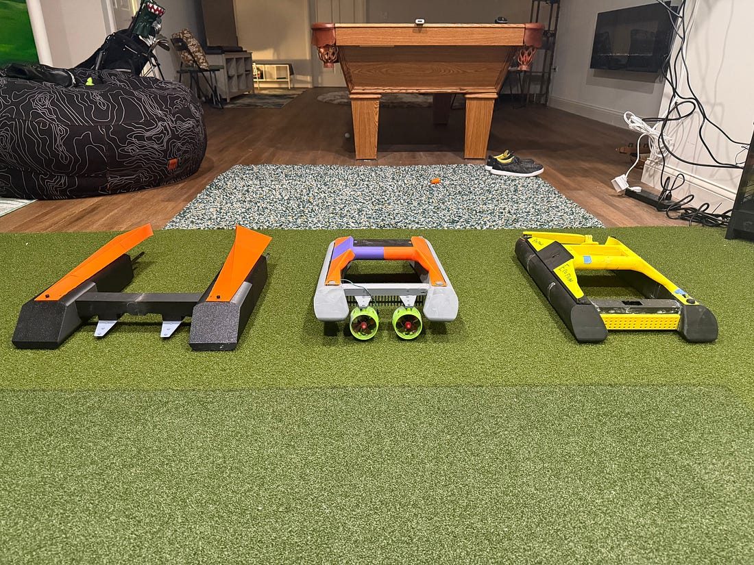 Three versions of the EcoFloat boat lined up on green turf in a basement: a black-and-orange hull on the left, a gray-and-purple one with two green thrusters in the middle, and a yellow one on the right.