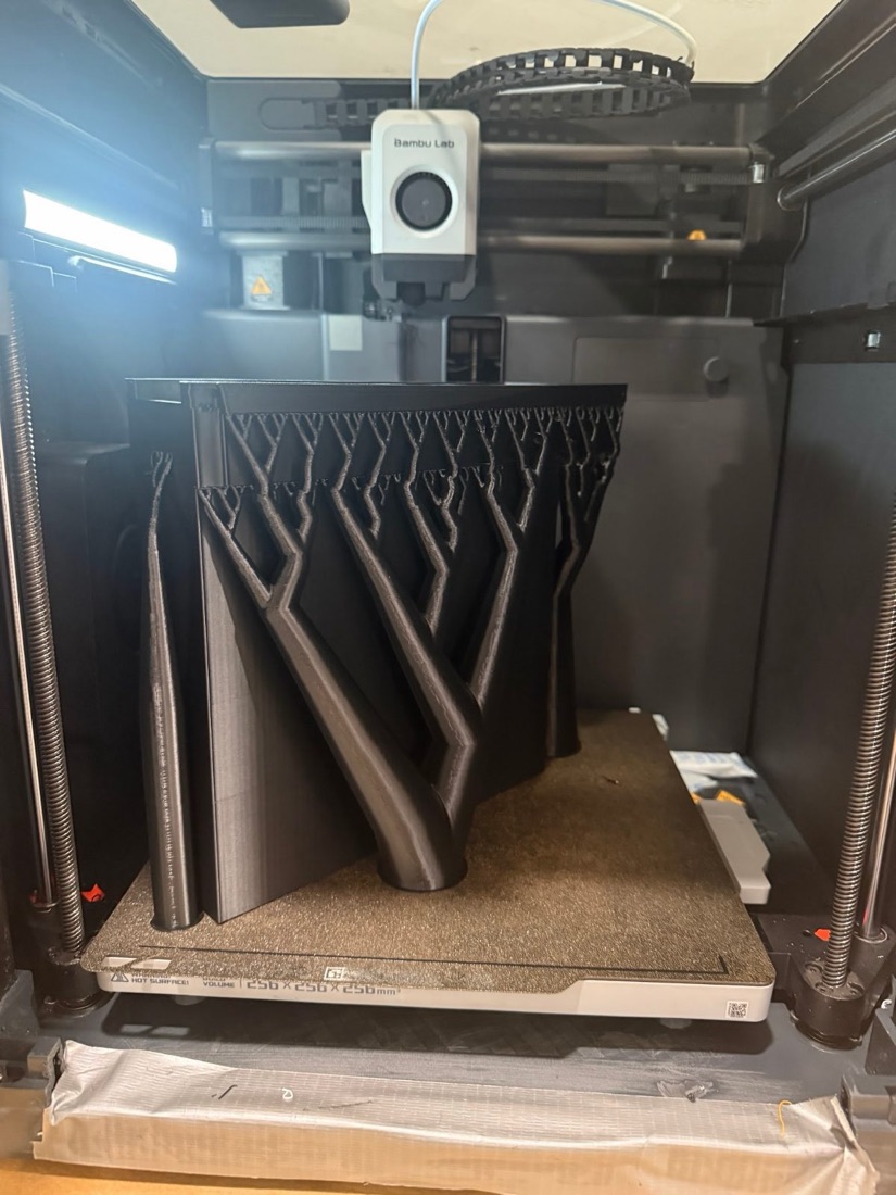 A large black part printing inside a Bambu Lab 3D printer, held up by branching tree-shaped supports.