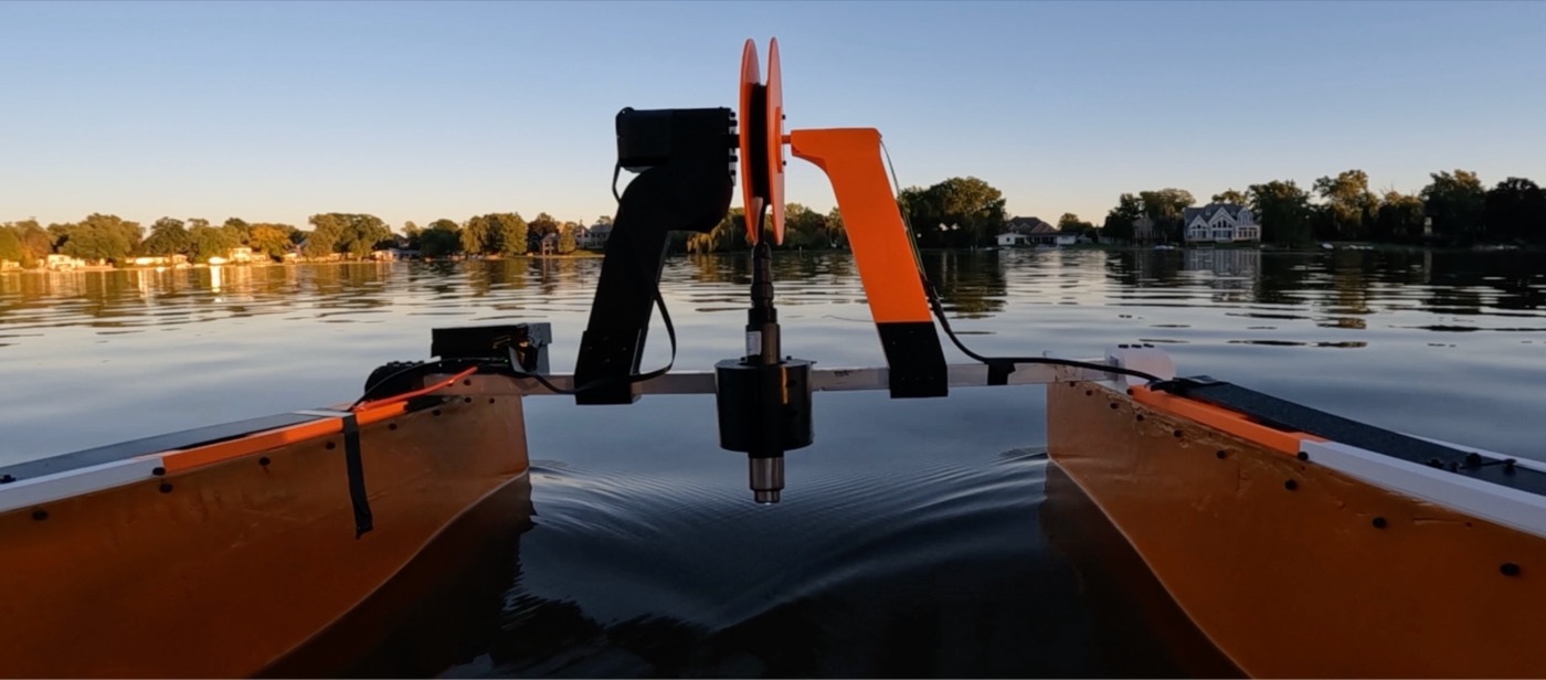View from the back of EcoFloat on a calm lake at sunset: an orange cable spool on a black and orange 3D-printed frame, with the oxygen probe hanging down toward the water between the two hulls.