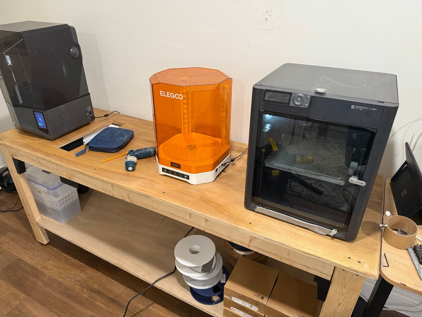 A wooden workbench holding a black resin printer, an orange Elegoo wash and cure station, and a Bambu Lab P1S printer, with filament spools on the shelf below.